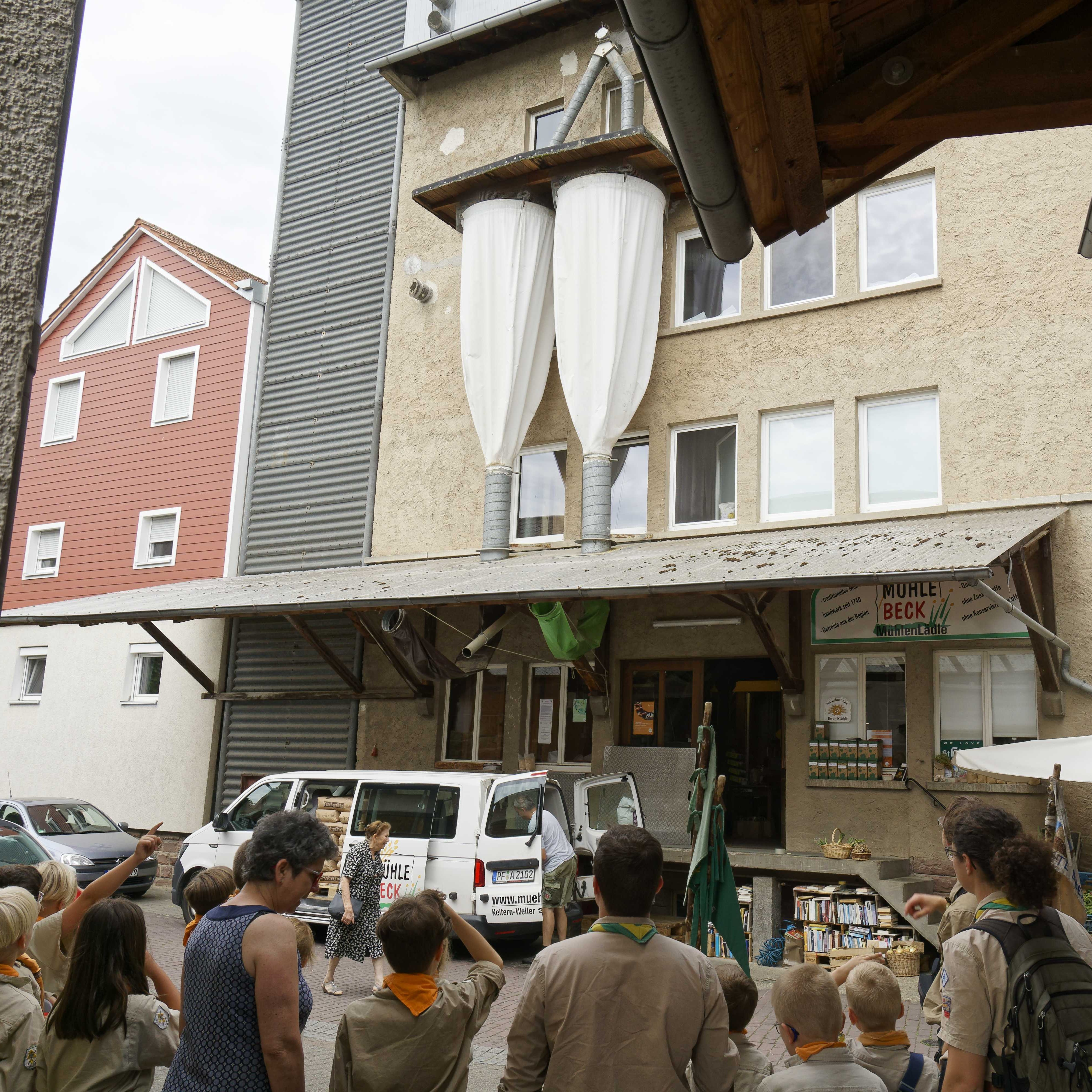 Vor der Mühle im Hof stehen die Jungen und Mädchen mit der Müllerin, während diese erzählt was es mit dem Äußeren der Mühle auf sich hat